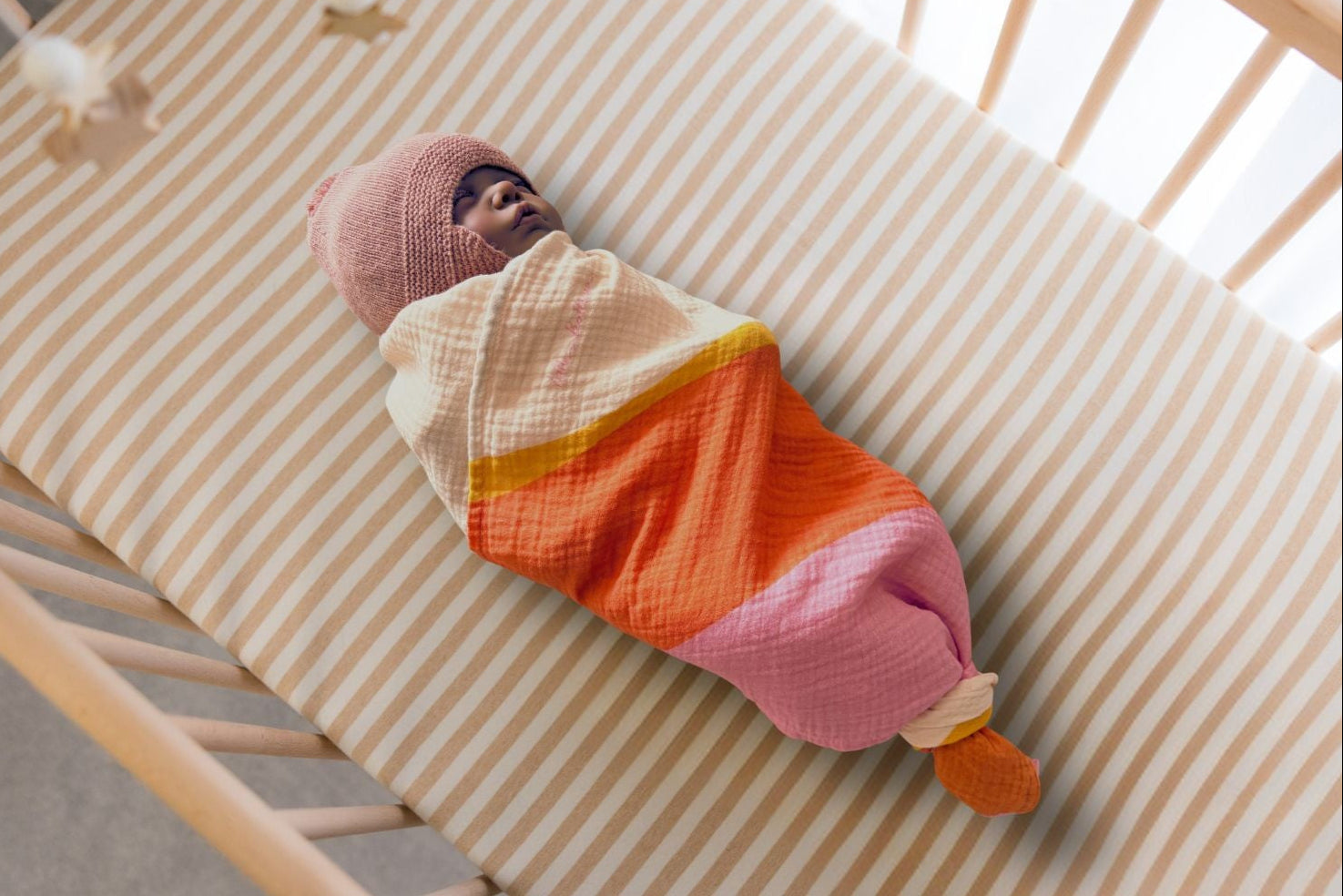 Baby swaddled in a colorful muslin blanket lying in a crib with striped bedding.
