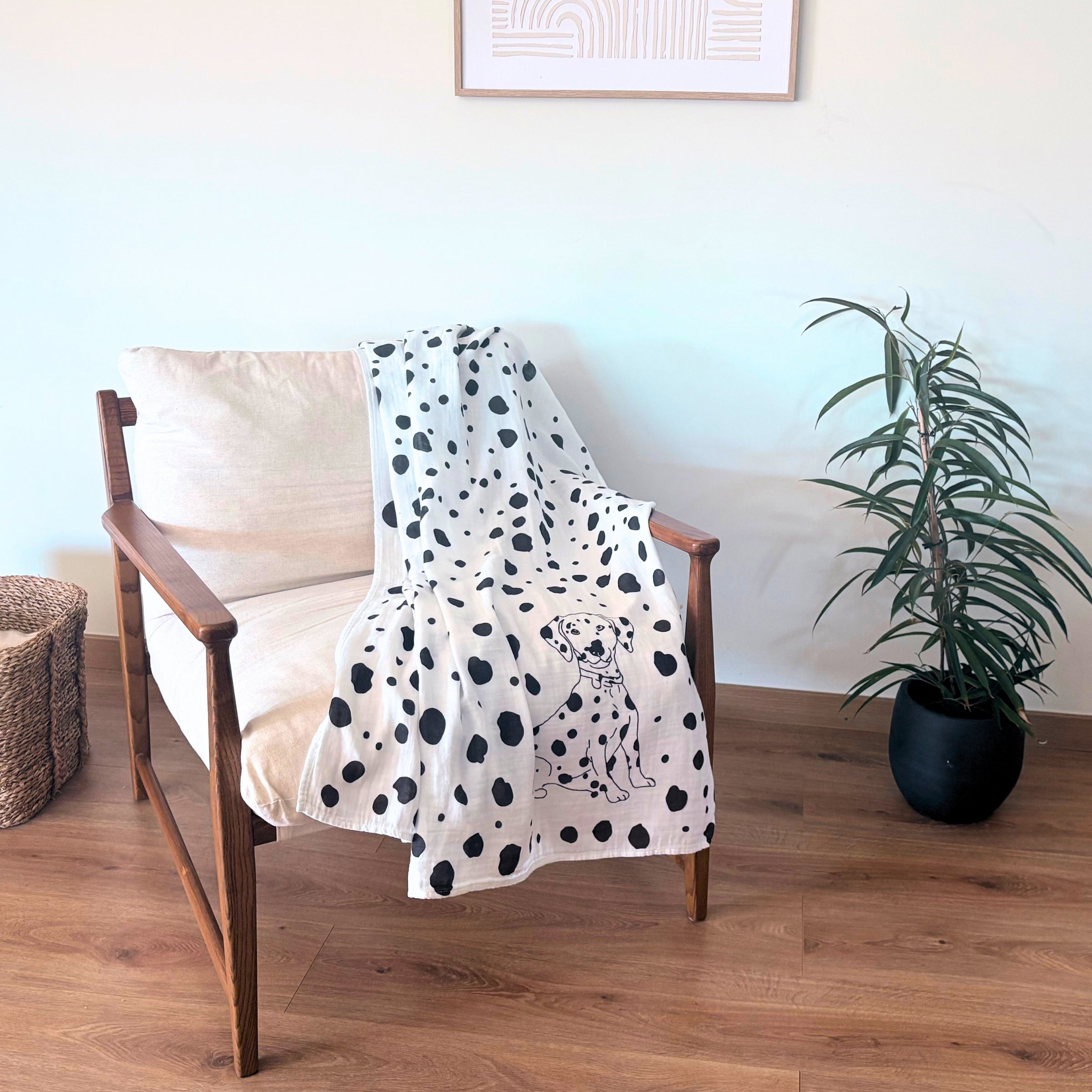 Modern chair with a Allure en Noir Dalmatian patterned Large Square blanket draped over it, next to a plant on a wooden floor.