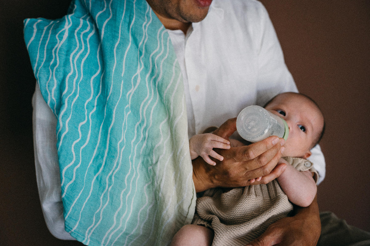 A man wearing a white shirt holds a baby dressed in a beige knit romper while feeding the baby with a clear bottle. A turquoise and white wavy-patterned muslin blanket is draped over his shoulder against a dark brown background.