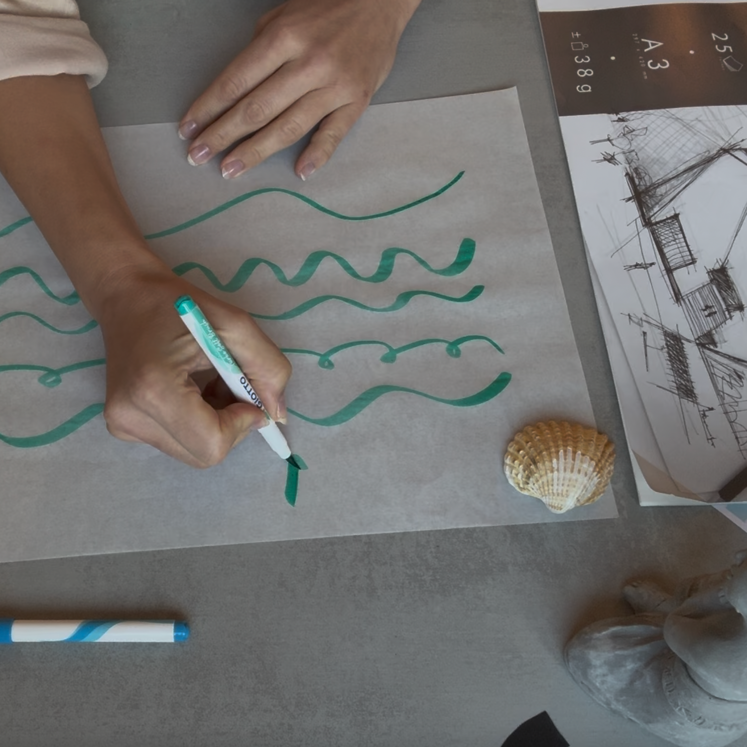 Person drawing wavy lines on a piece of paper with a marker, surrounded by seashells and stationery.