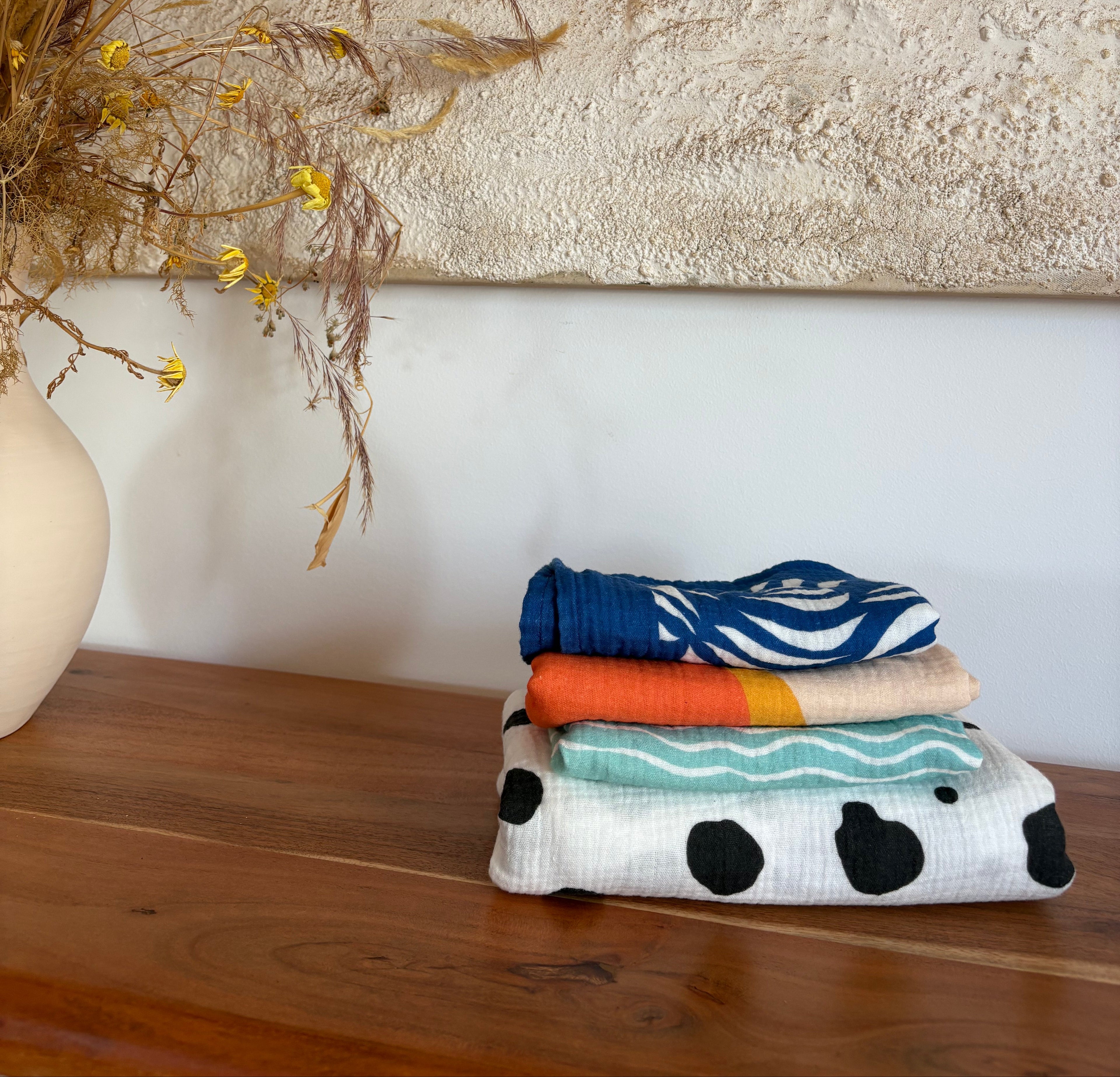 Product image: 4 designs of the organic muslin swaddles on a stylish wooden table, natural light.