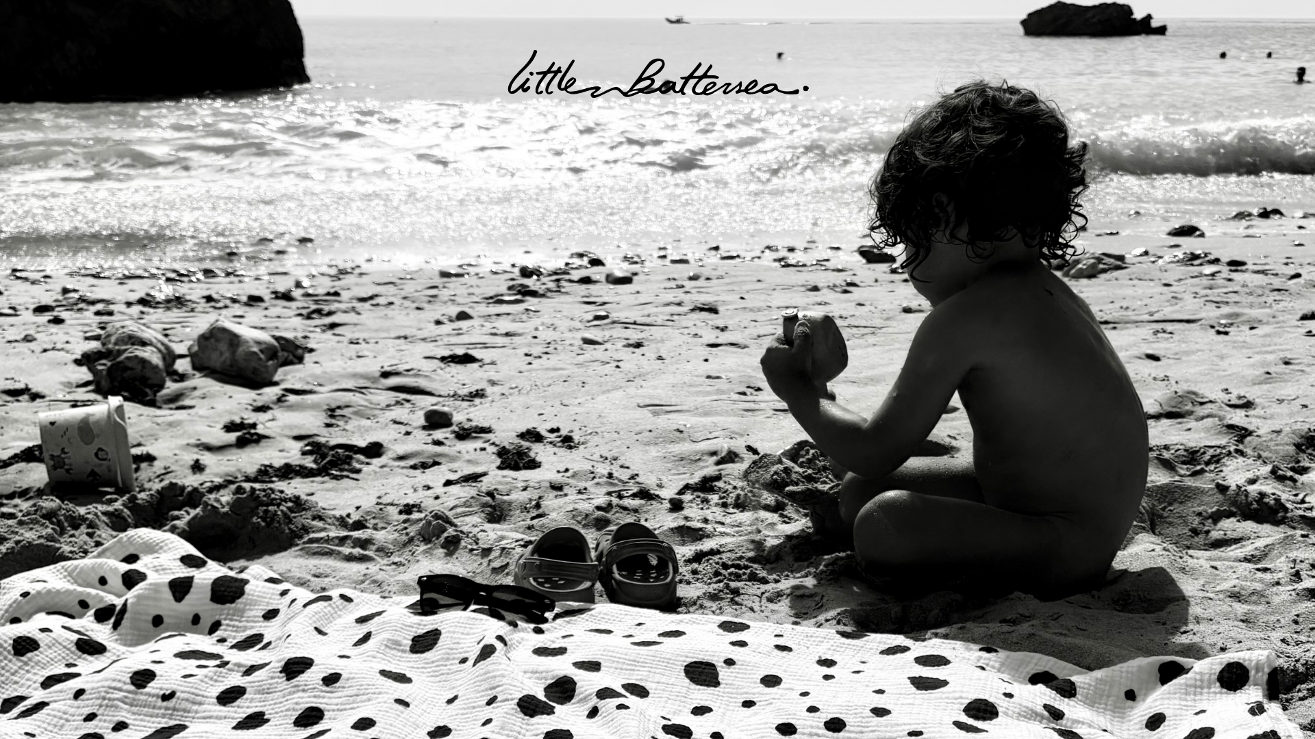 A child playing on a beach with a Little Battersea muslin Allure en Noir spread out, embodying the blend of parenthood and personal style.