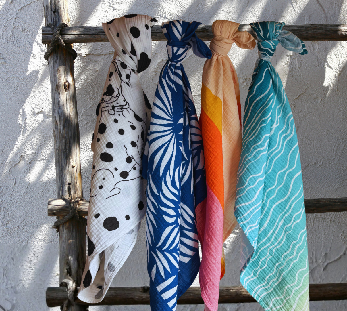 Five stylish muslins hanging on a wooden ladder against a white wall. Fünf stilvolle Musselin-Tücher hängen an einer Holzleiter vor einer weißen Wand.