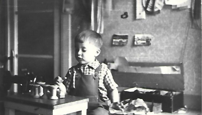 The son of Erich Schwarze, a young boy intently engaging with tailoring tools at a table, hinting at a lifelong dedication to design.
